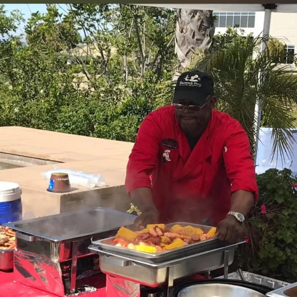 A chef in a red shirt and black cap serves steaming shrimp, corn, and sausage from a buffet.