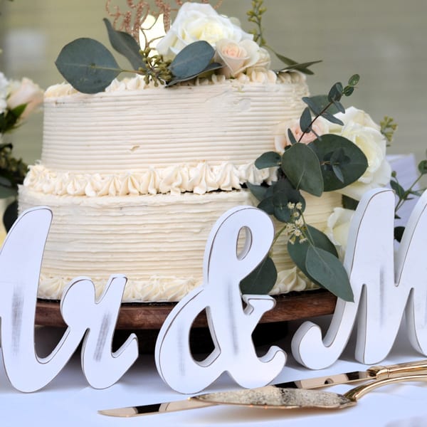 Two-tiered white wedding cake with white roses, eucalyptus, and "Mr & Mrs" sign.