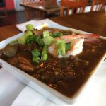 Dark seafood gumbo topped with chopped green onion and shrimp in a white square bowl set on a dark wood table.
